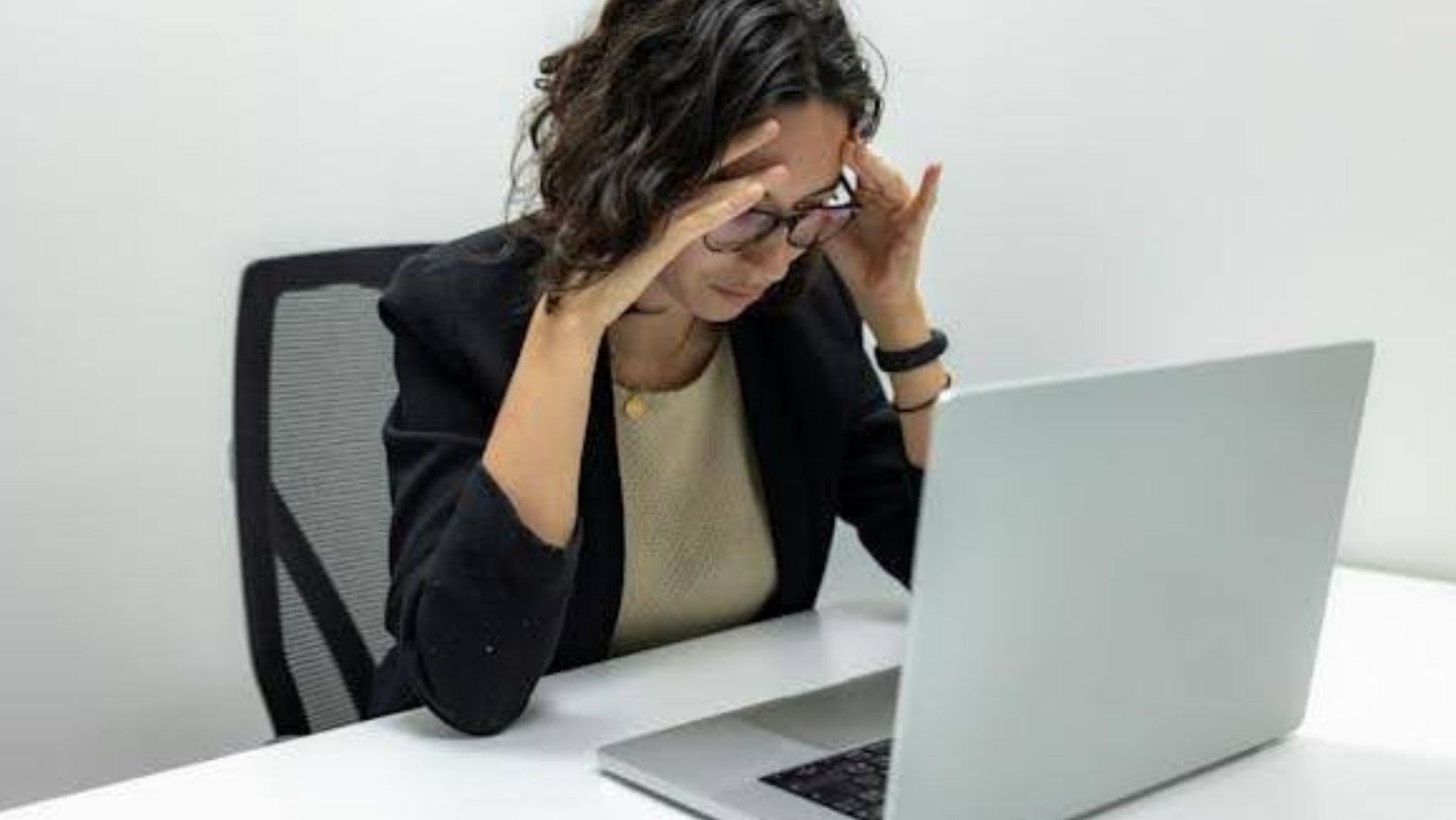A woman in business attire sitting in front of a laptop and holding her hands to her head.